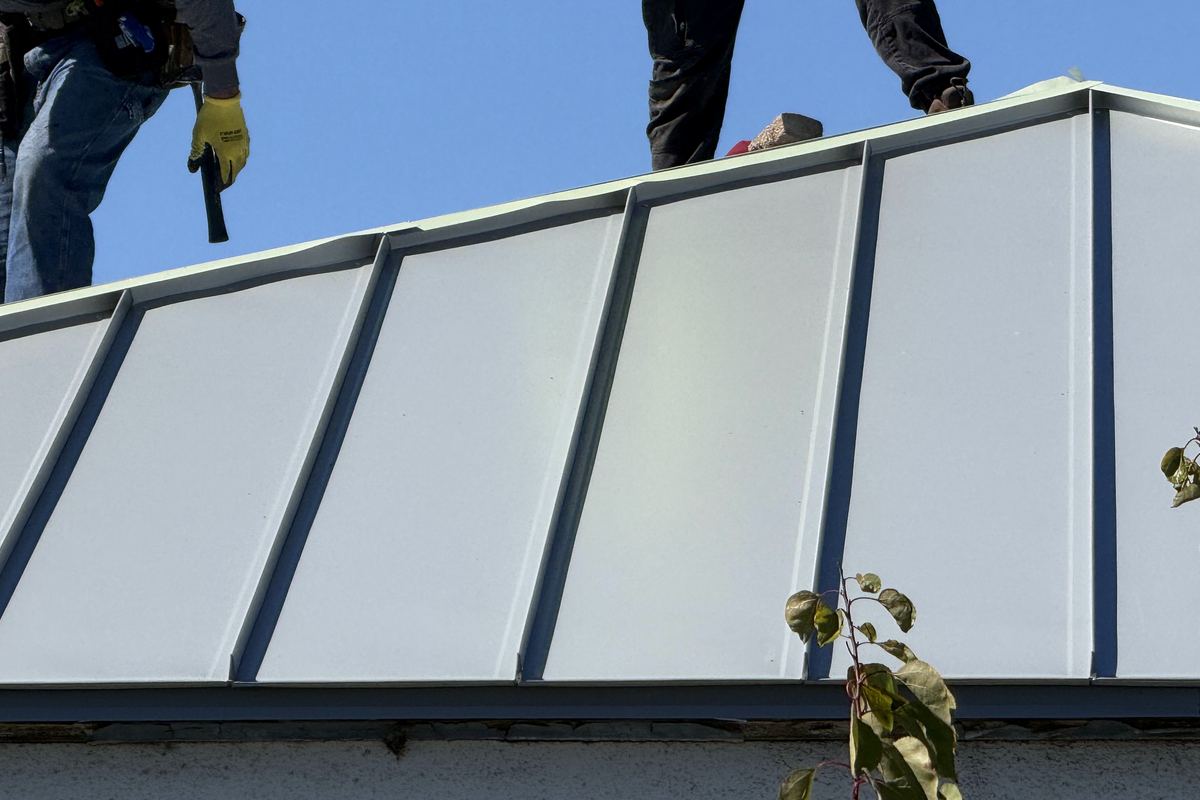 Daylight view of two roofers working at a standing-seam metal roof edge.